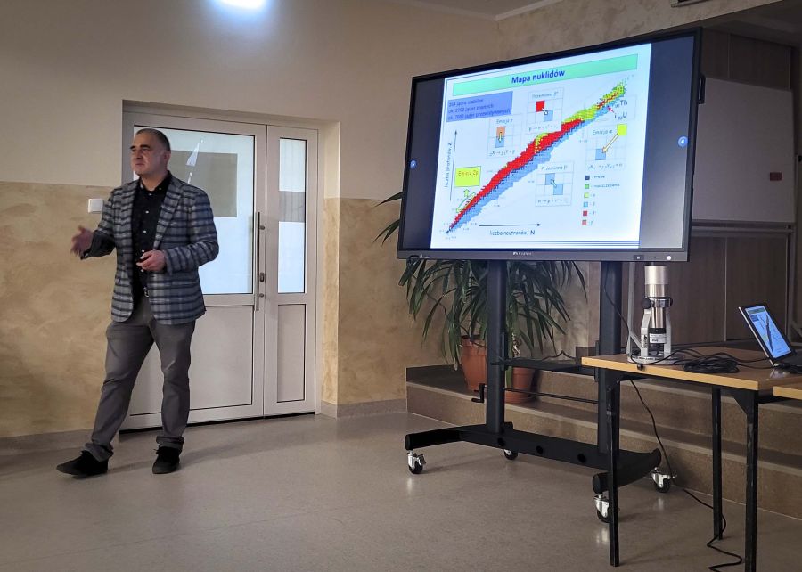 mężczyzna w marynarce cos opowiada obok ekran z wyświetlonym obrazem