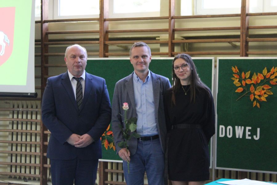 dwóch męzczyzn i dziewczyna w okularach stoją obok siebie, mężczyzna w środku trzyma różę