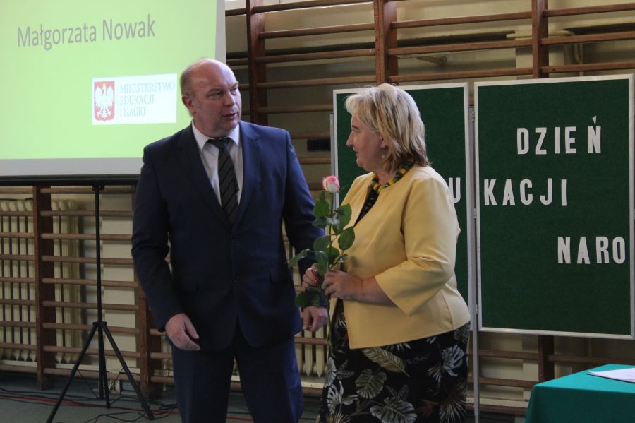 mężczyzna gratuluje kobiecie, kobieta trzyma kwiatek w tle tablica z napisem i ekran z wyświetlanym obrazem