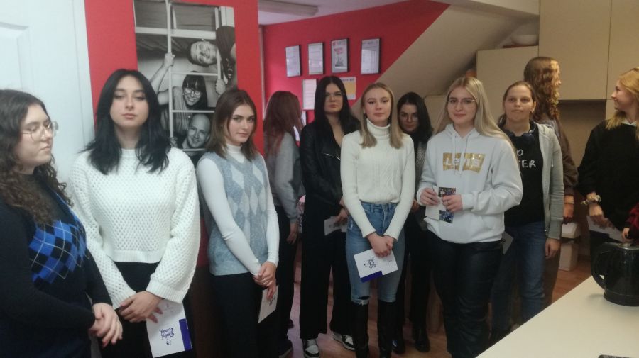 Uczennice stoją w pomieszczeniu z ulotkami w ręce i pozują do zdjęcia. W tle widać zdjęcia osób oraz dyplomy.