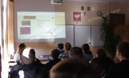 uczniowie widoczni od tyłu siedzą w ławkach w klasie oglądają pokazywaną na ekranie prezentację