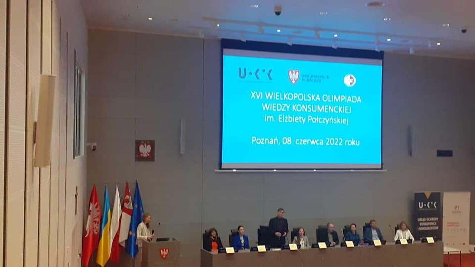 Jury konkursu oraz prelegenci siedzący przy stole prezydialnym. W tle rzutnik z informacjami o olimpiadzie.