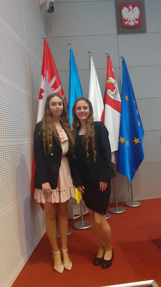 Uśmiechnięte dwie uczennice biorące udział w konkursie. W tle flagi.
