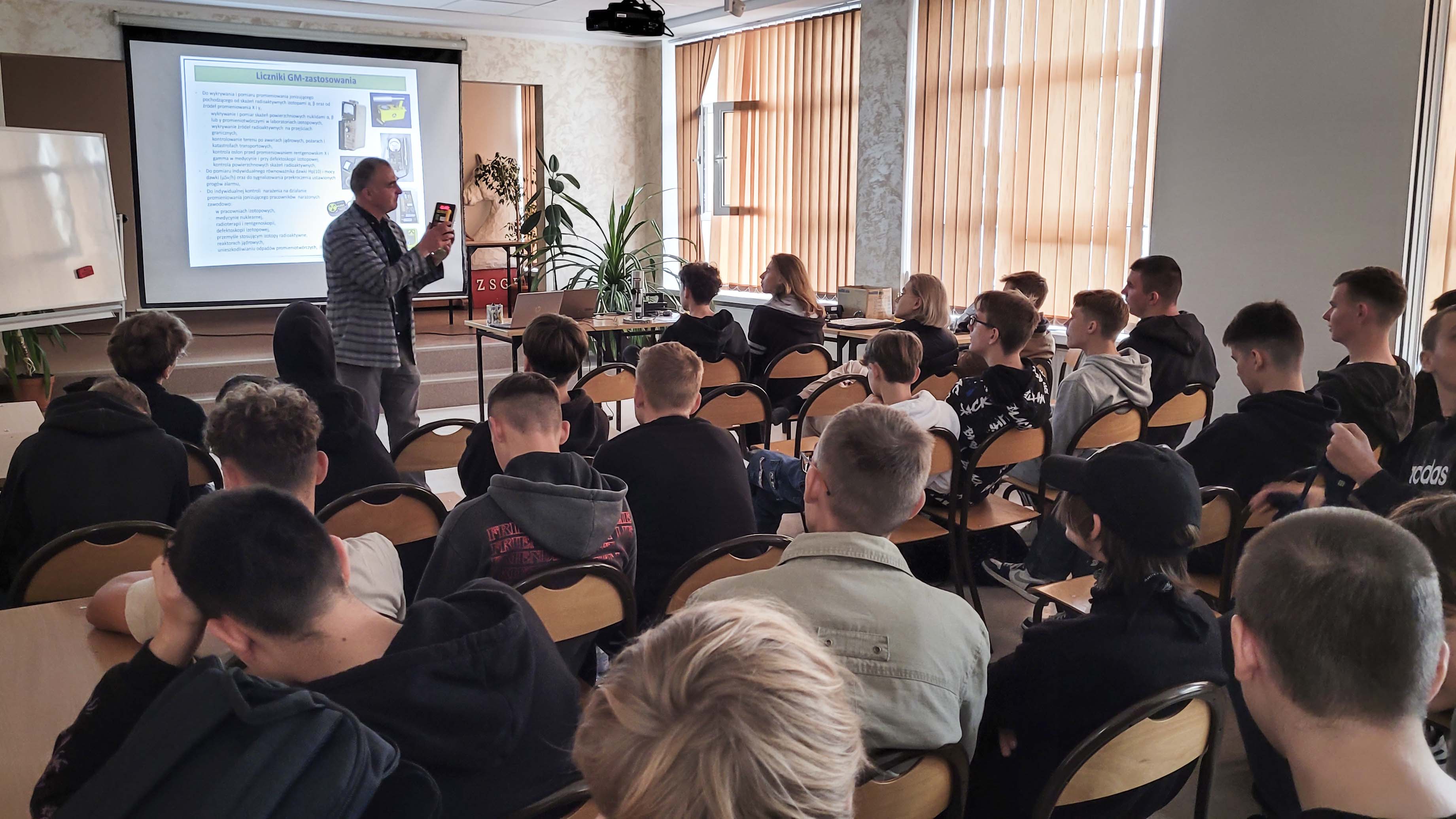grupa uczniów siedzi na krzesłach tyłem do aparatu przed nimi stoi mężczyzna w marynarce coś opowiada
