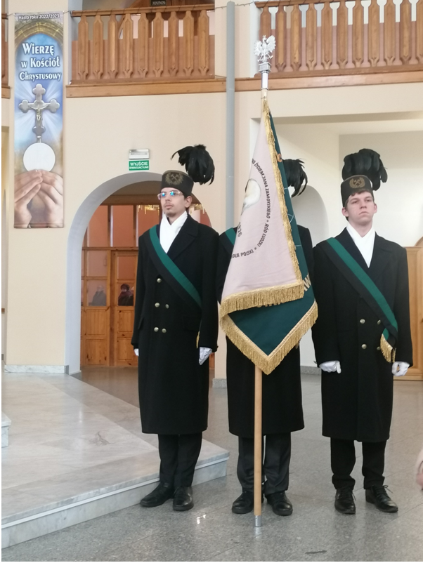 Na zdjęciu poczet sztandarowy ZSGE w Koninie.  Stoi na tle ściany , od strony kaplicy adoracji Najświętszego Sakramentu w kościele NMP Królowej Polski w Koninie. Poczet sztandarowy tworzą    trzej uczniowie ubrani w czarne płaszcze , białe szale, na głowa